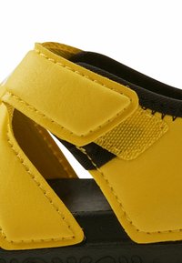 Sandales synthétiques jaunes avec des accents noirs, dotées d'une fermeture à bande Velcro, d'une surface texturée et d'une semelle en caoutchouc moulé.
