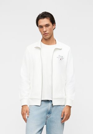 Jonge man met donker haar die een wit Tommy Hilfiger jack met rits en lichtblauwe spijkerbroek draagt, staand tegen een effen achtergrond.