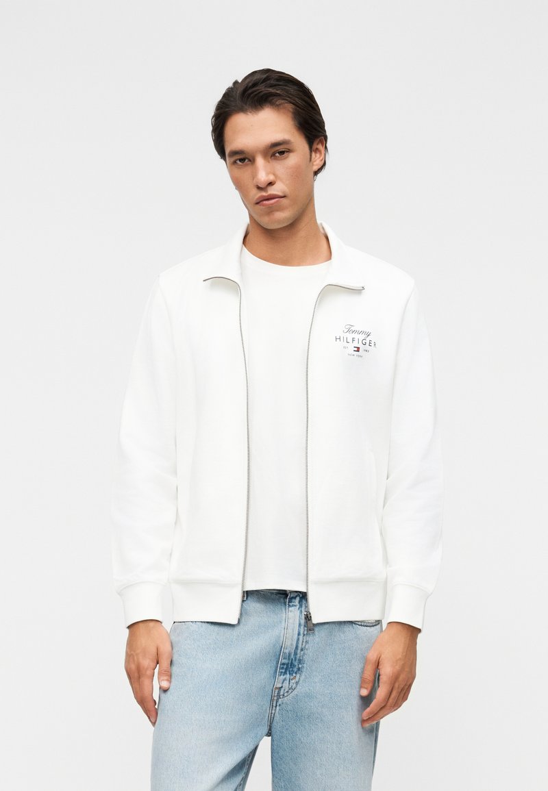 Jeune homme aux cheveux foncés portant une veste blanche zippée Tommy Hilfiger et un jean bleu clair, debout devant un fond uni.