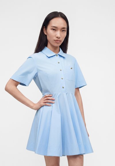 Γυναίκα που φοράει ένα κοντομάνικο ανοιχτό μπλε φόρεμα με κολάρο, χρυσά κουμπιά και πλισέ φούστα, στέκεται με το ένα χέρι στη μέση της.