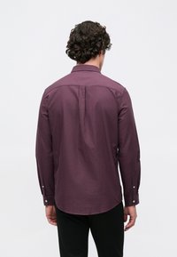 Homme aux cheveux bouclés et foncés portant une chemise violette à manches longues et un pantalon noir, vu de dos sur un fond blanc uni.