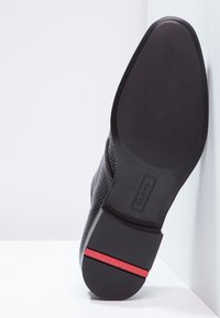 Semelle de chaussure noire avec une prise texturée, présentant une finition lisse et un accent rouge près du talon. Le design est épuré et moderne.