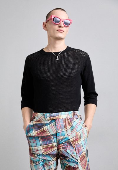 Schwarzer, Netzstoff, langärmeliger Pullover kombiniert mit mehrfarbigen, karierten Hochbundhosen und pinken, eckigen Sonnenbrillen. Silberne Anhängerkette.