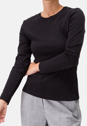 Person wearing a fitted black long-sleeve shirt and gray pants, adjusting the left sleeve with their right hand.