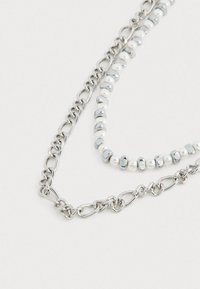 Collier en chaîne métallique argenté comportant des perles alternant entre brillantes et texturées, y compris des perles blanches, sur un fond clair.