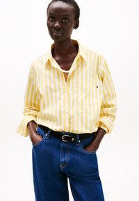 Camicia a righe gialle e bianche con taschino, abbinata a jeans in denim blu a vita alta e una cintura nera.