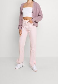 Kvinna som bär ljusrosa högmidjade jeans med sidoklipp, vit tube-top, mauve stickad kavaj och vita sneakers.