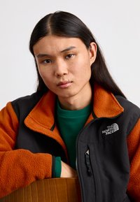 Giacca in pile nera e arancione con zip, con il logo North Face. Indossata sopra un maglione verde, mette in evidenza colori e trame contrastanti.