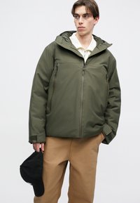 Maslinasto zelena izolirana jakna s kapuljačom, s dvije džepove na zip, podešavajućim manžetama i glatkom teksturom. Nosiva s bež hlačama.