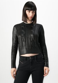 Femme avec des cheveux noirs lisses portant un pull en maille noir texturé et un jean noir taille haute sur fond blanc.