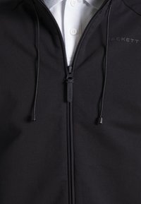 Giacca nera con zip e cappuccio, realizzata in tessuto liscio. Presenta un logo sul lato sinistro e lacci al collo. Sotto, è visibile una camicia bianca con colletto.