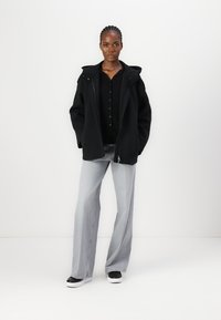 Black, zip-up jacket with a hood, paired with a black button-up shirt and light grey wide-leg jeans. Black and white sneakers complete the look.