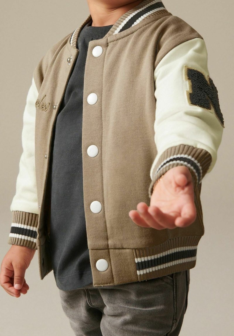 Enfant portant une veste universitaire beige et blanche avec un t-shirt noir et un pantalon gris, tendant la main droite vers l'avant avec la paume ouverte.