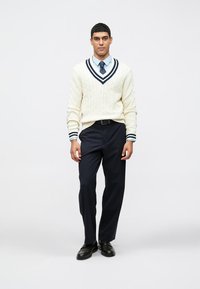 Άνδρας που φοράει κρεμ πουλόβερ με πλέξη καλωδίου και ναυτικές ρίγες, ανοιχτό μπλε πουκάμισο, ναυτικό παπιγιόν, μαύρο παντελόνι και μαύρα παπούτσια, στέκεται μπροστά σε λευκό φόντο.