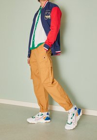 Marinblå och röd collegemantel med broderad patch, vit skjorta med gröna accenter, cargo-byxor i ljusbrunt och flerfärgade sneakers.