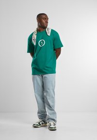 Camiseta verde con un gran logo blanco en forma de "1", jeans de mezclilla de lavadero claro y zapatillas verdes y blancas. Una bufanda floral está drapeada alrededor del cuello.