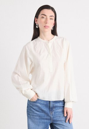Una persona che indossa una blusa a strisce di colore chiaro e jeans blu è in piedi contro uno sfondo bianco, mostrando un look di moda casual.