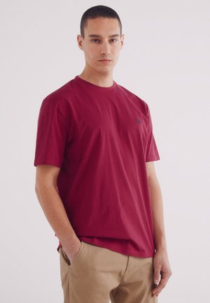 Bordeauxfarbiges Baumwoll-T-Shirt mit Rundhalsausschnitt, kurzen Ärmeln und einem dezenten gestickten Logo auf der Brust, kombiniert mit hellbeigen Hosen.