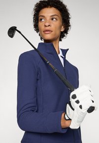 Sötétkék, texturált, cipzáras dzseki, testhezálló fazonban, fehér, galléros ing párosításában. Golfütőt tart a kezében, fehér kesztyűben, fekete részletekkel.