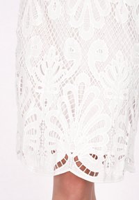 Abito bianco di pizzo con motivi floreali e fogliari, con dettagli intagliati intricati e orlo a scallop. Tessuto leggero e semi-trasparente.