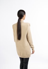 Pull beige en maille avec col montant et épaules étendues, présentant une coupe décontractée et des poignets côtelés, porté avec un pantalon noir ajusté.