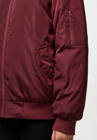Burgundy puffer jacket with zippered sleeve pocket, ribbed cuffs, and hem. Smooth, shiny fabric; tailored fit, minimal seams.