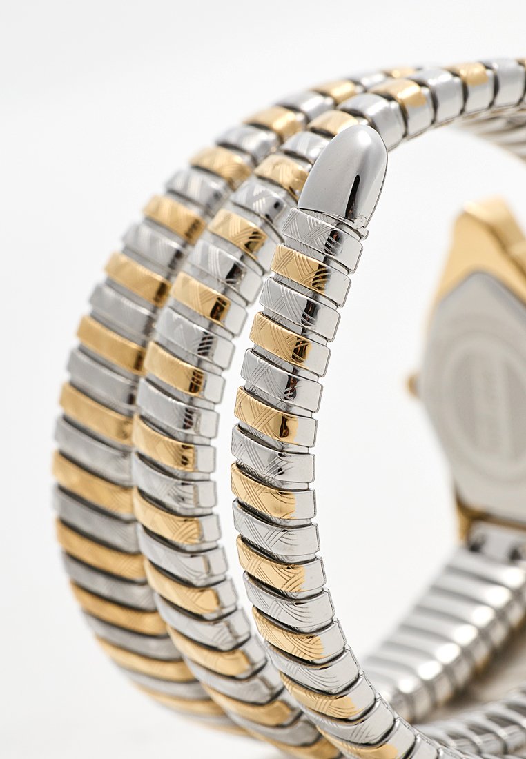 Orologio in metallo argento e oro con un bracciale flessibile che presenta anelli alternati texture e un'estremità arrotondata lucida. Quadrante dell'orologio esagonale distintivo.