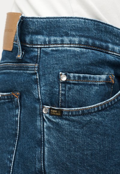 Zbliżenie na tylną kieszeń niebieskich dżinsów z metalowymi nitami i małą metką marki Tiger of Sweden na białym tle koszuli.