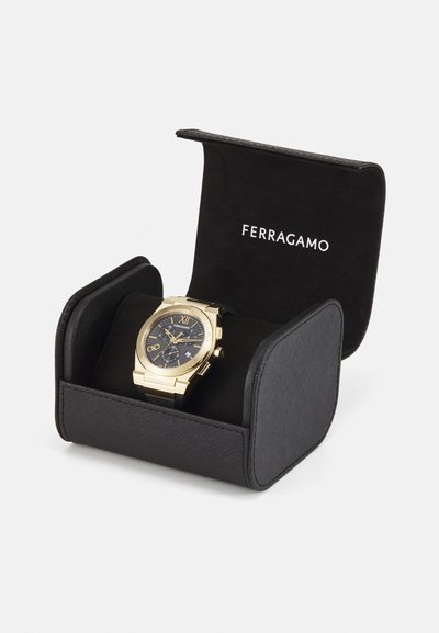 Gold-tone watch with a navy dial, Roman numerals, date feature, and black leather strap, displayed in a black fabric-lined box with logo.