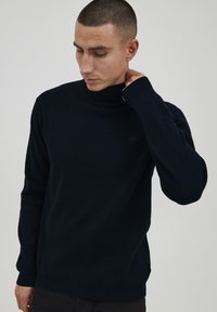 Pulover de culoare bleumarin cu guler înalt, fabricat dintr-un material tricotat moale. Prezintă manșete și tiv cu nervuri, cu un detaliu subtil al logo-ului pe piept.