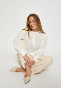 Ilga balta bluzė su dryžiais, su mandarinų apykakle ir mygtukų uždarymu, derinama su šviesiai bežinėmis plačiomis kelnėmis ir nude spalvos smailiomis batais.