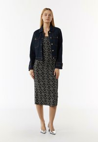 Denimjacke mit goldenen Hardware-Elementen über einem schwarzen Kleid mit einem weißen abstrakten Muster, kombiniert mit glänzenden silbernen High Heels.