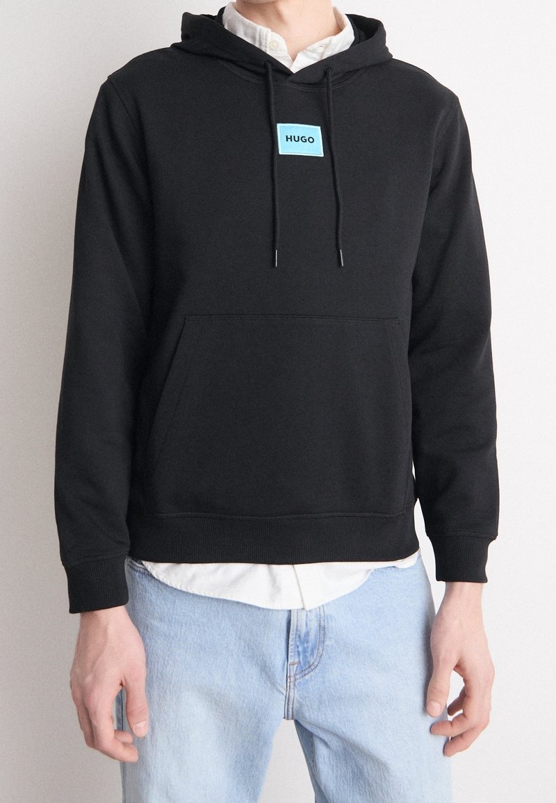 Sudadera negra con capucha, bolsillo canguro, cordones ajustables y un parche de marca azul claro en el pecho. Material de mezcla de algodón, ajuste relajado.