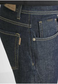 Donkerblauwe spijkerbroek met gele stiksels. Kenmerken zijn onder andere meerdere zakken en een klein metalen accent. Het materiaal lijkt duurzaam en met een structuur.