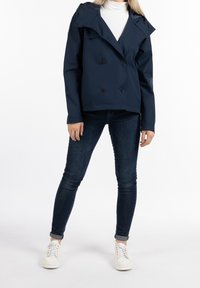 Chaqueta navy de doble botonadura con grandes solapas, hecha de tela suave. Combinada con jeans oscuros ajustados y zapatillas blancas, de pie sobre un fondo blanco.