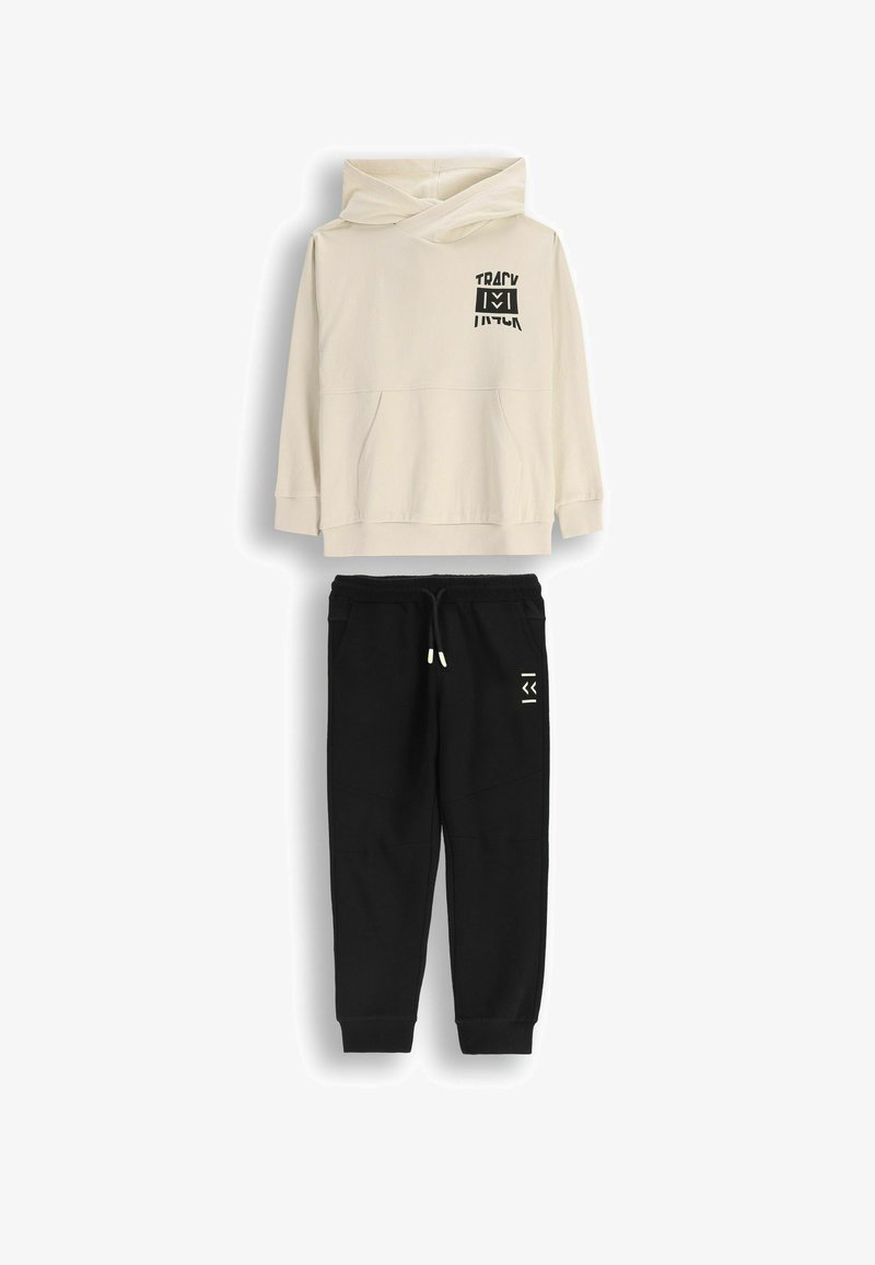 Jasny beżowy hoodie z przednią kieszenią kangurką oraz czarnym nadrukiem graficznym, w zestawieniu z czarnymi spodniami dresowymi z bocznymi kieszeniami i logo.