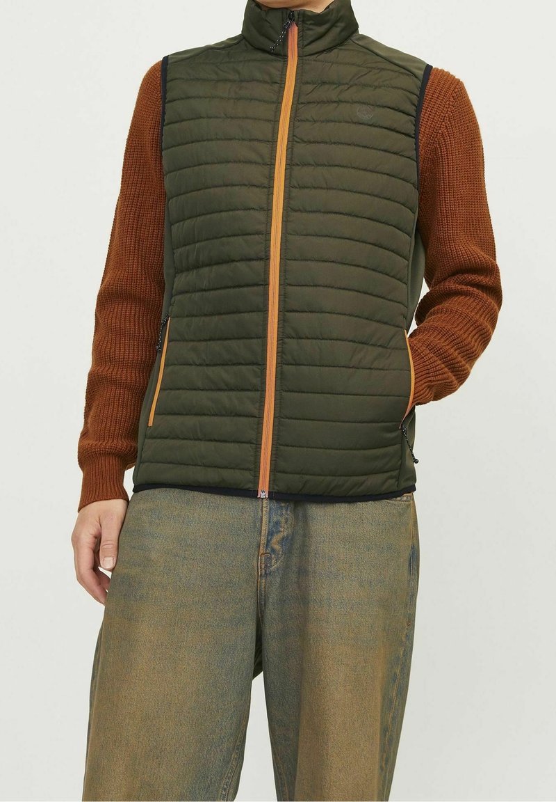 Zielona ocieplana kamizelka z pomarańczowym zamkiem błyskawicznym, wysoki kołnierz, boczne kieszenie i kontrastowe brązowe dzianinowe rękawy, w połączeniu z luźnymi niebieskimi dżinsami.