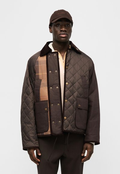 Moški, oblečen v rjavo prešito jakno z rjavim karo podlogom, rjavo kapo in temne hlače, obrnjene proti kameri na enobarvni svetli podlagi.