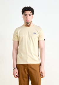 Beige, rövid ujjú póló, pamutból készült, kicsi kék logóval a bal mellrészén és texturált felülettel. Barna nadrággal párosítva.
