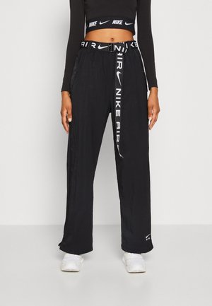 Osoba ubrana w czarne spodnie Nike Air z dopasowanym paskiem i czarny krótki top, w białych trampkach, stojąca na jednolitym tle.