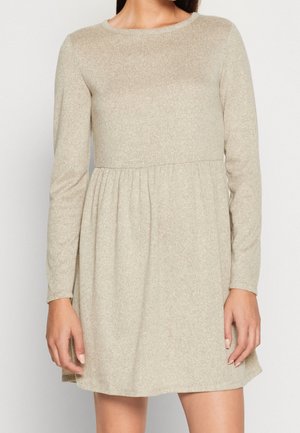 Frau trägt ein beiges Kleid mit langen Ärmeln, das über das Knie reicht, mit einem taillierten Oberteil und einem gerafften Bund vor einem einfarbigen Hintergrund.