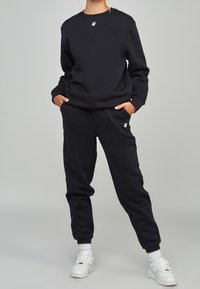Sudadera negra y pantalones de chándal a juego en material suave. Presenta puños acanalados y cinturilla elástica. Pequeño logo en el pecho y en los pantalones. Calzado blanco.