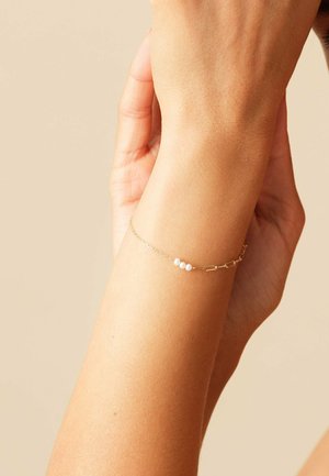 Handgelenk mit zartem Goldkettchen-Armband, das drei kleine weiße Perlen auf einem neutralen beigen Hintergrund zeigt.