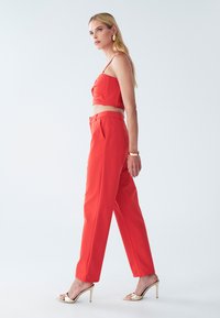 Crvena kratka majica s teksturiranim detaljima i krojenim hlačama viskog struka. Zlate sandale s visokom petom i dodaci dopunjuju odjeću.