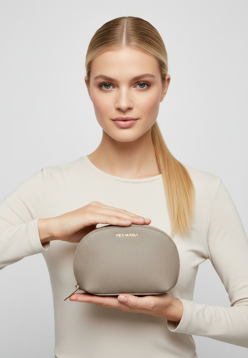 Beige textuur cosmetica pouch met een ronde vorm, met "HEY MARLY" in goud gebosst, vastgehouden door een persoon tegen een lichte achtergrond.