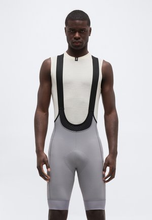 Muškarac u bijeloj majici bez rukava s crnim bretelem i sivim biciklističkim hlačicama s umetnutim jastučićima stoji ispred jednobojne pozadine.