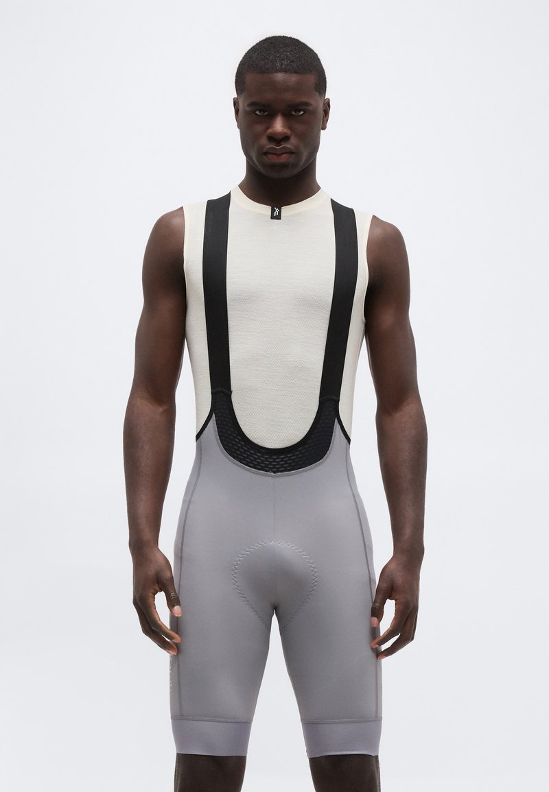 Άνδρας που φοράει λευκό αμάνικο τοπ με μαύρους τιράντες και γκρι επενδεδυμένα ποδηλατικά σορτς με τιράντες, στέκεται μπροστά σε απλό φόντο.