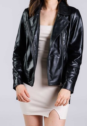 Vrouw draagt een zwarte leren jas, losgeritst, over een lichtbeige mouwloze jurk met een kleine voorsplit.