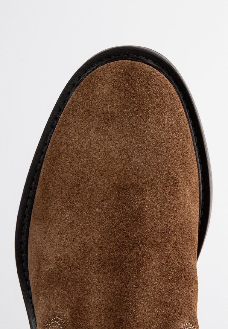 Brązowy zamszowy but z okrągłym noskiem, czarną podeszwą i kontrastowym przeszyciem wzdłuż krawędzi, prezentujący miękką teksturę.