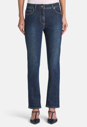 Femme portant un jean droit bleu foncé et des escarpins à lanières en T bleu marine à bout pointu, debout devant un fond uni.